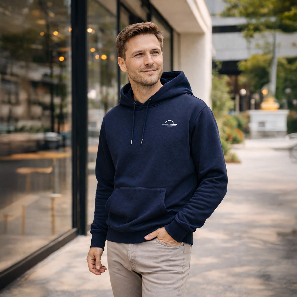 Man wearing a navy blue organic cotton hoodie with a subtle sunset logo, standing outdoors near a building.