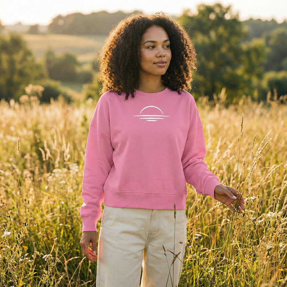 Woman wearing a, organic pink cotton sweatshirt with a minimal sunset design in a field during sunset